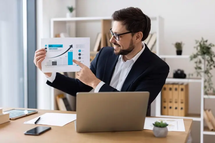 A businessman sitting at a desk with a laptop, holding and pointing to a printed chart showing upward growth during a presentation in a modern office setting.