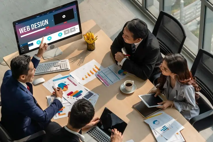 Four business professionals sitting around a conference table reviewing web design analytics on a desktop screen, with charts and graphs spread across the table in a modern office setting.